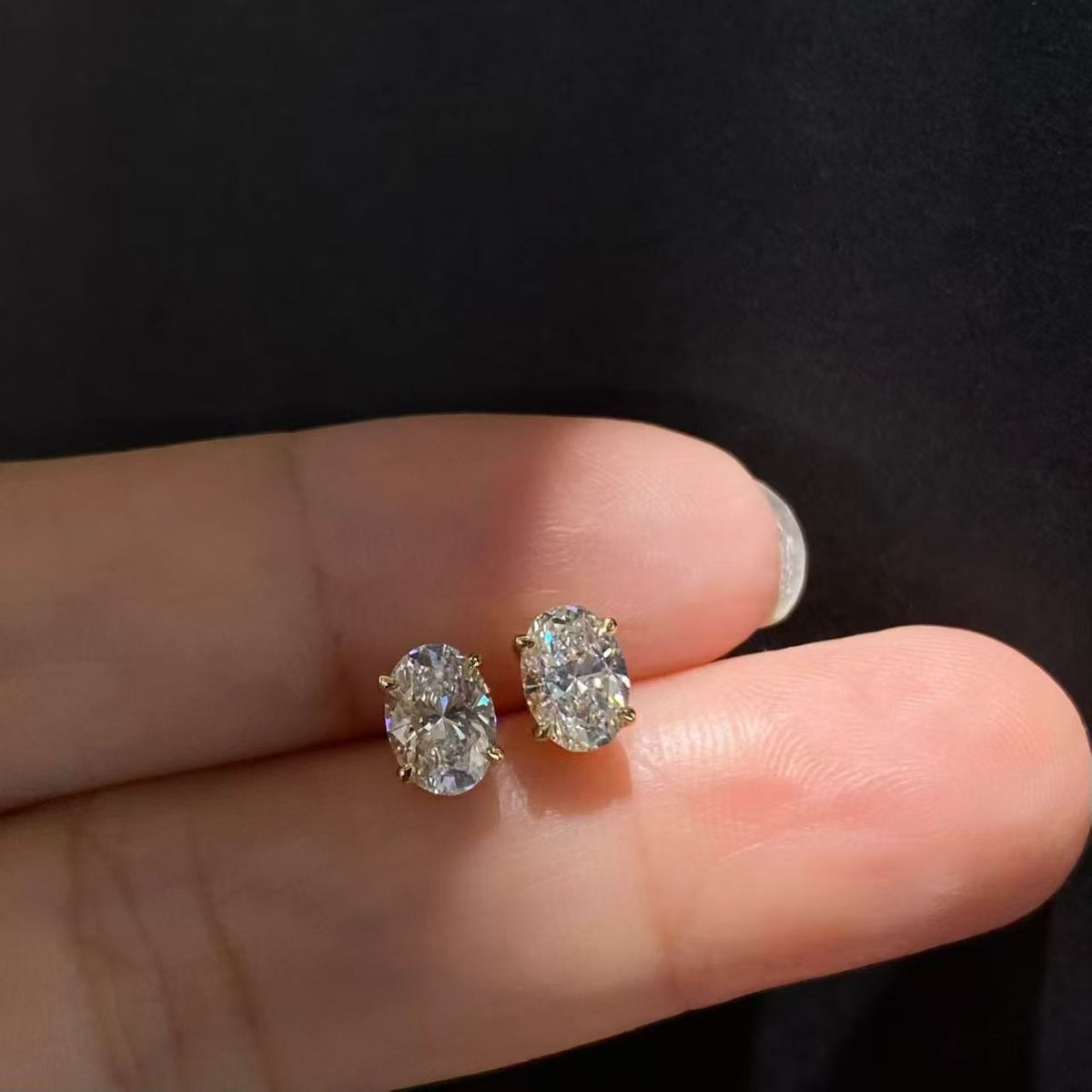 Oval solitaire lab diamond stud earrings held on fingertips - front view