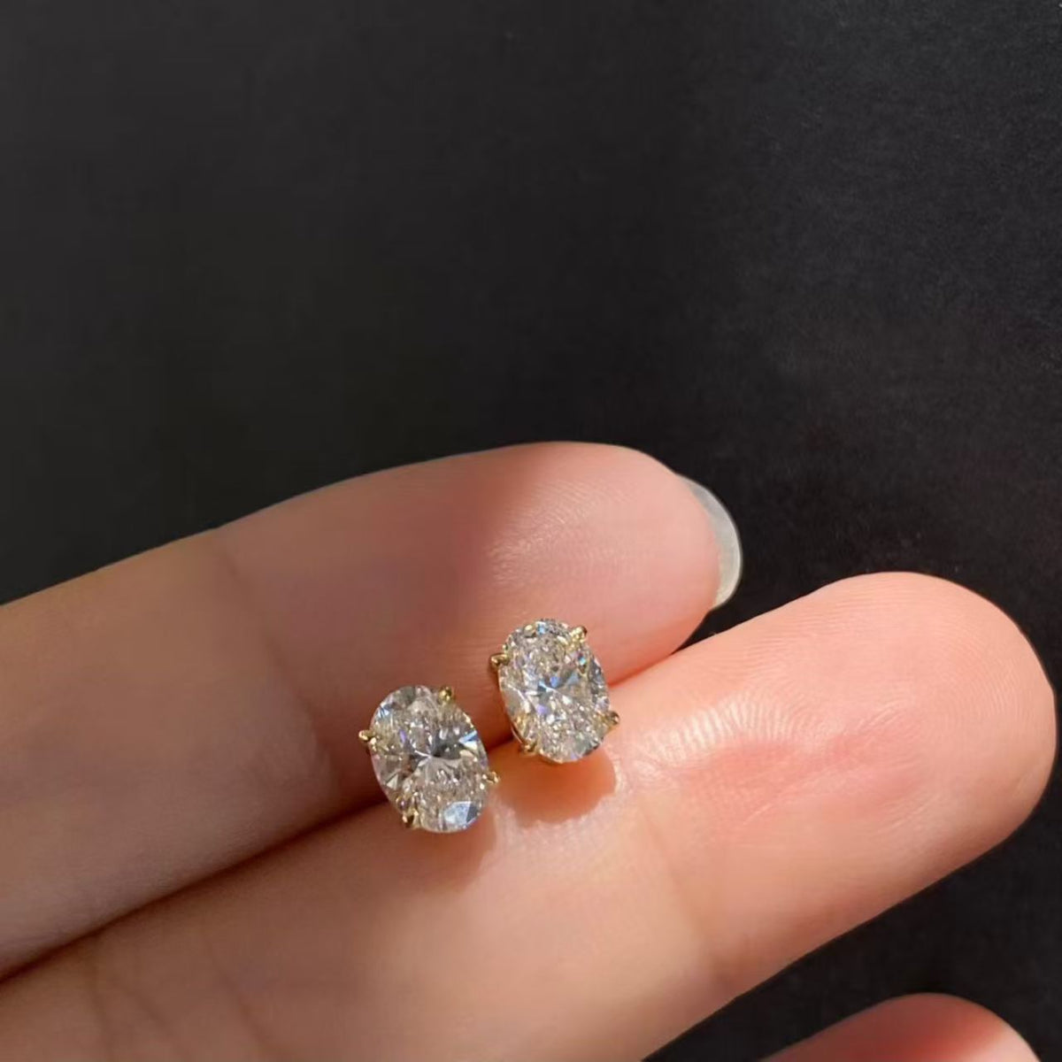 Oval solitaire lab diamond stud earrings held on fingertips - angle view