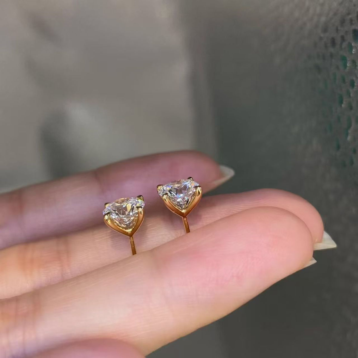Heart and round lab diamond stud earrings held on fingertips in 18K yellow gold